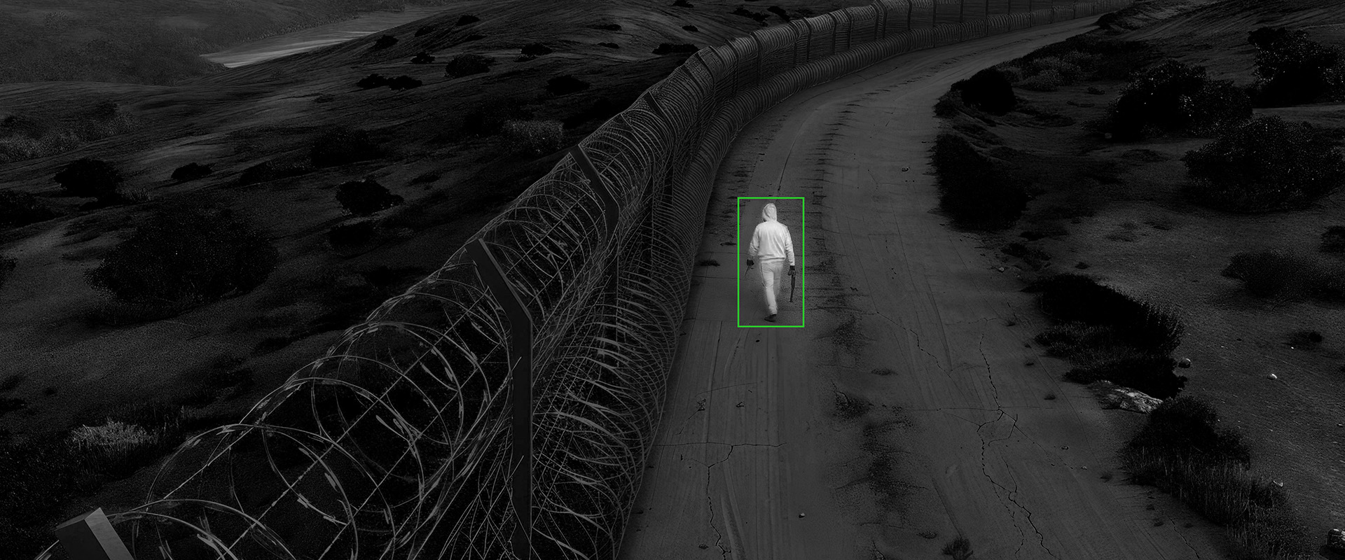 Eine Person im Hoodie wird von einer Wärmebild-Überwachungskamera neben einem Maschendrahtzaun erkannt. 